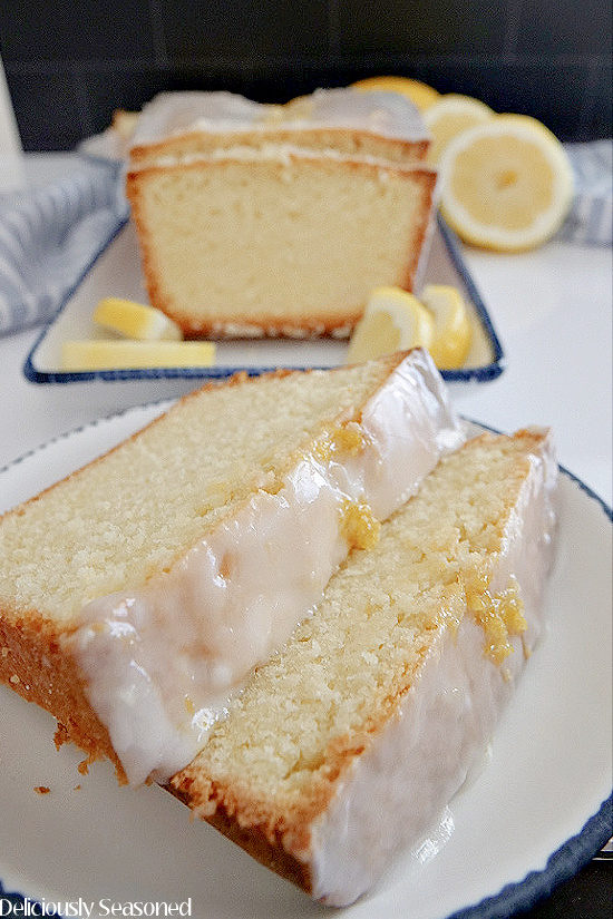 Two slices of lemon pound cake on a white plate with the loaf cake in the background along with lemon slices.