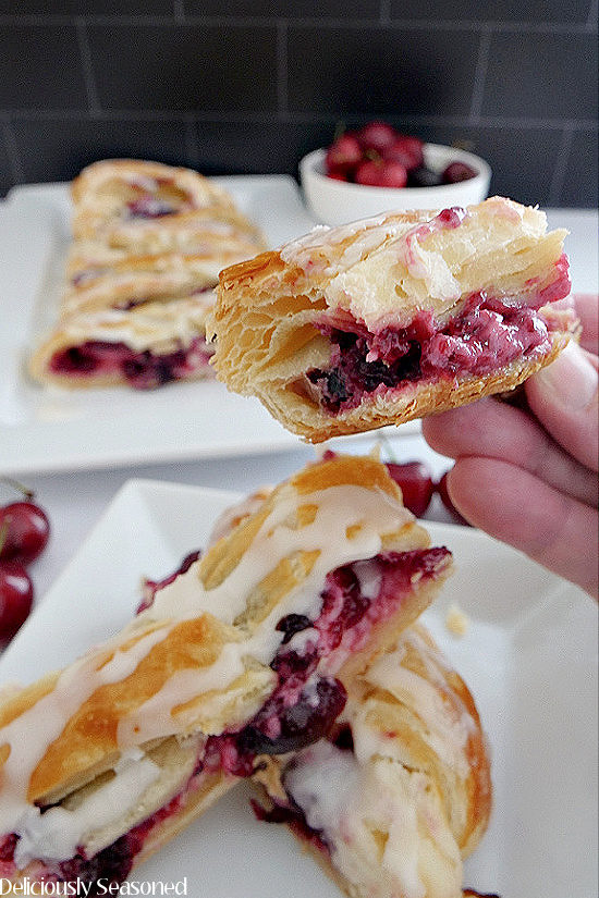 A slice of Cherry Cream Cheese Danish with a bite taken out being held up to show the filling, with cherries and more slices in the background.