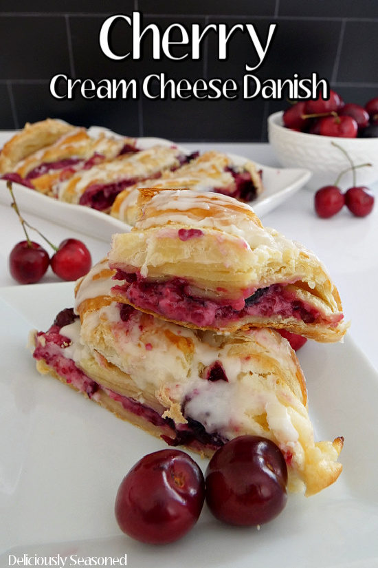 Two slices of Cherry Cream Cheese Danish on a white plate stacked on top of each other with a white oblong plate in the background with more slices of the Danish on it.