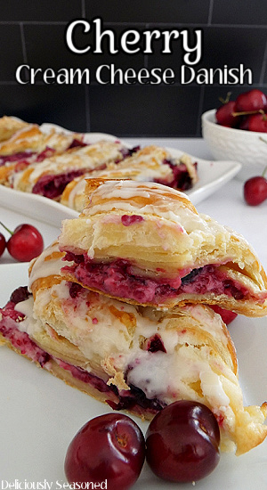 Two slices of Cherry Cream Cheese Danish on a white plate with two cherries in front and more slices and cherries in the background.