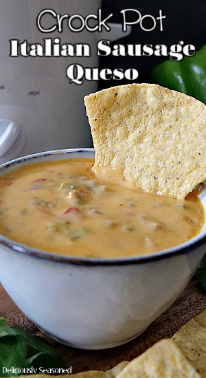 A small grey bowl filled with queso with Italian sausage and a tortilla chip placed in the queso, also a small white crock pot in the background.