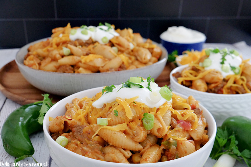 Two white bowls and a grey bowl filled with taco pasta with a small blue bowl of sour cream in the background, along with jalapenos, cilantro and green onions.