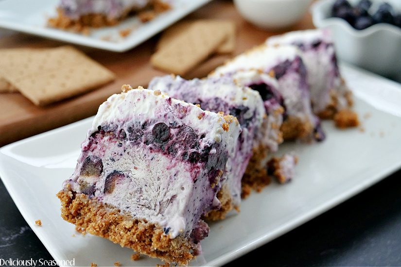 A white plate with 5 thin slices of frozen blueberry dessert with blueberries, graham crackers and more dessert in the background.