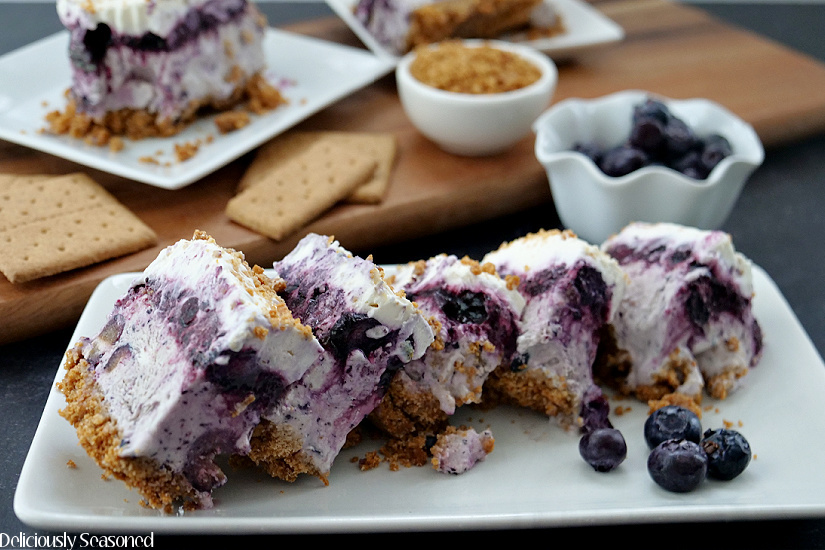 A white oblong plate with 5 slices of frozen blueberry dessert placed on it with fresh blueberries in the background along with more frozen dessert and graham crackers.