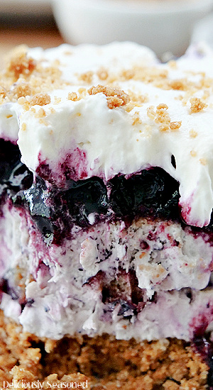 A super close of photo of a piece of frozen blueberry dessert showing the graham cracker crust, blueberry cream cheese filling, blueberry topping and then topped with homemade whipped cream.