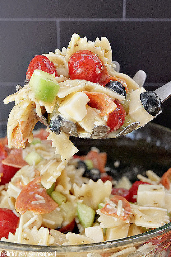 A spoonful of Bow Tie Pasta over a large bowl of Bow Tie Pasta showing the sliced black olives, halved grape tomatoes, ¼ pepperoni pieces, diced cucumbers and chunks of mozzarella cheese. 