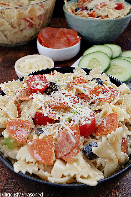 Bow Tie Pasta Salad in a black bowl, showing the Farfalle noodles, pepperoni, grape tomatoes, sliced black olives, mozzarella cheese cubes, and grated parmesan cheese.