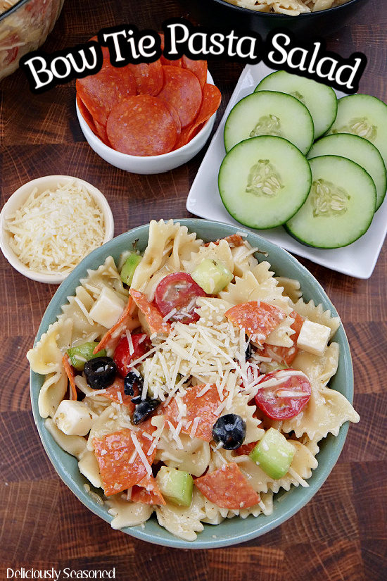 An overhead picture of Bow Tie Pasta Salad in a blue bowl, showing the halved grape tomatoes, sliced olives, chunks of mozzarella cheese, pepperoni pieces, and diced cucumbers, topped with grated parmesan cheese. 