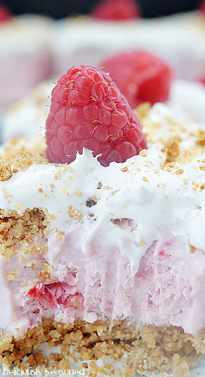 A close up photo of a slice of frozen raspberry dessert showing the graham cracker crust, the raspberry creamy filling and the whipped topping and a raspberry on top.
