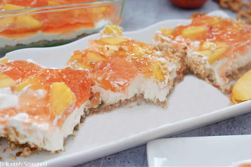 A long white plate with 3 slices of peach pretzel salad on it, with fresh peaches and pretzels in the background.