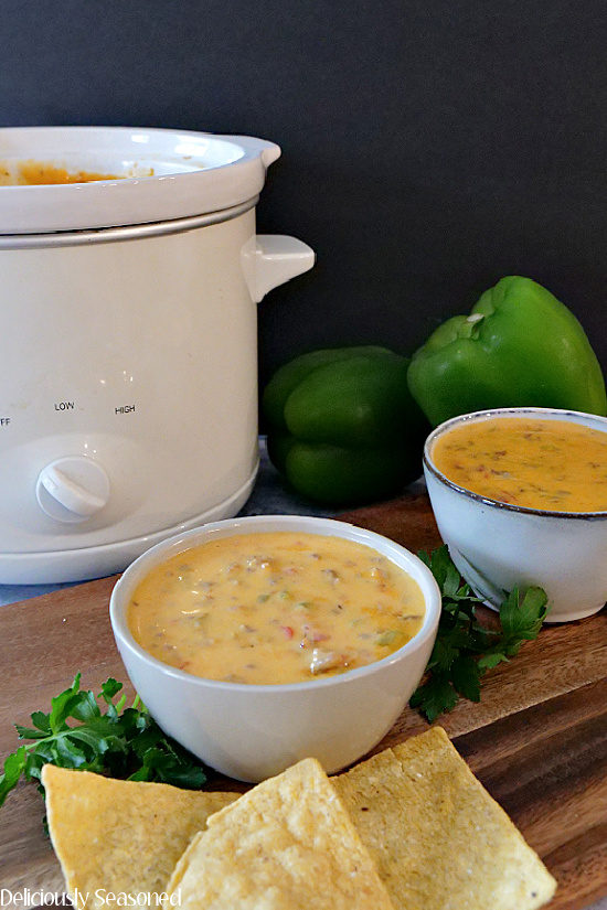 Two small bowls filled with crock pot queso with Italian sausage with a small white crock pot in the background with two bell peppers, all sitting on a wooden display board.