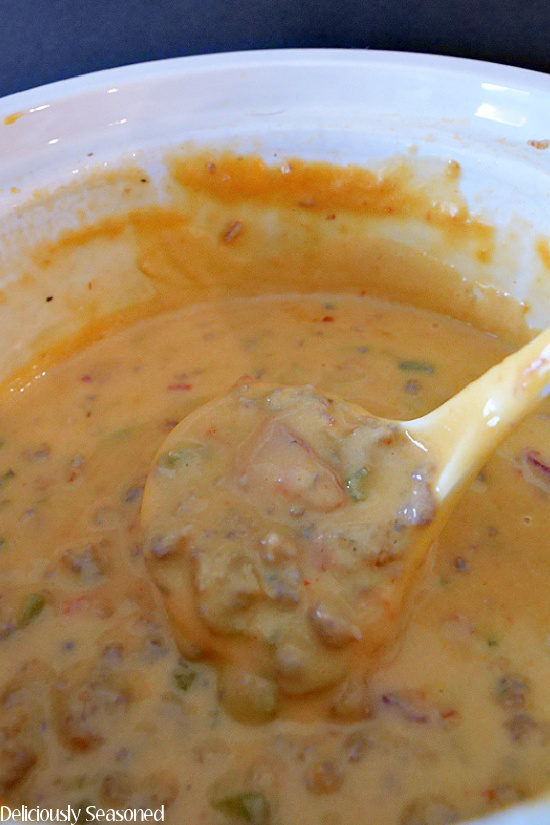 Looking down into a small white crock pot at the queso with Italian sausage and a small ladle scooping up so queso.
