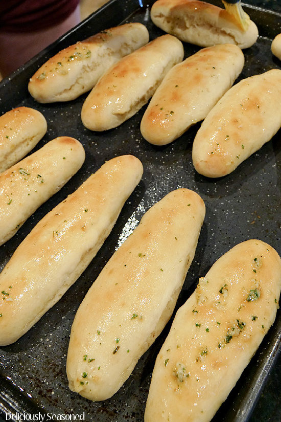A dark colored baking sheet with 11 breadsticks on it being topped with a melted butter mixture.