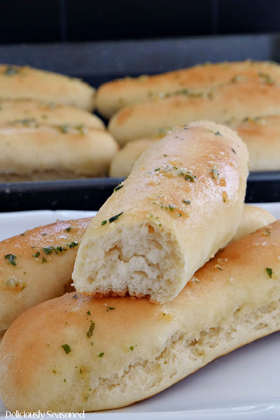 A close up photo of three breadsticks on a white plate with a bite taken out of one of them, and the baking sheet with more breadsticks in the background.