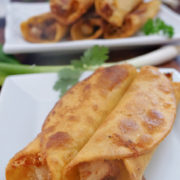 Two chicken taquitos on a white plate with another white plate with 6 taquitos on it in the background.