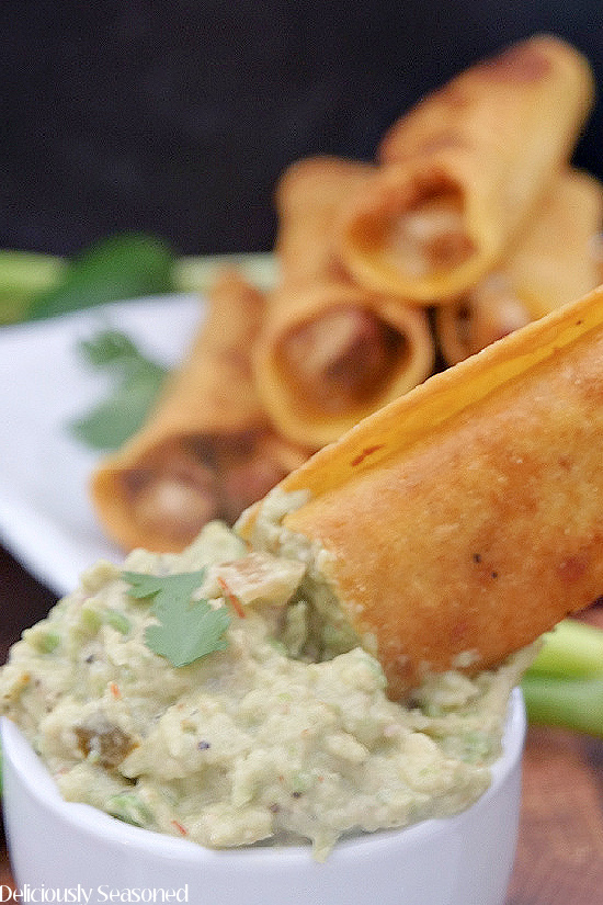 A chicken taquito being dipped into a white bowl filled with guacamole with more taquitos in the background.