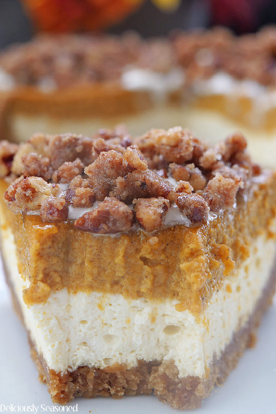 A close up photo of a slice of pumpkin cheesecake showing the layers of cheesecake, pumpkin pie on top of a graham cracker crust and topped with candied pecans.