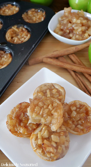 Five apple pie bites on a white plate with cinnamon sticks, green apples, small white bowl with apple pie filling and a mini muffin pan with more bites in it.