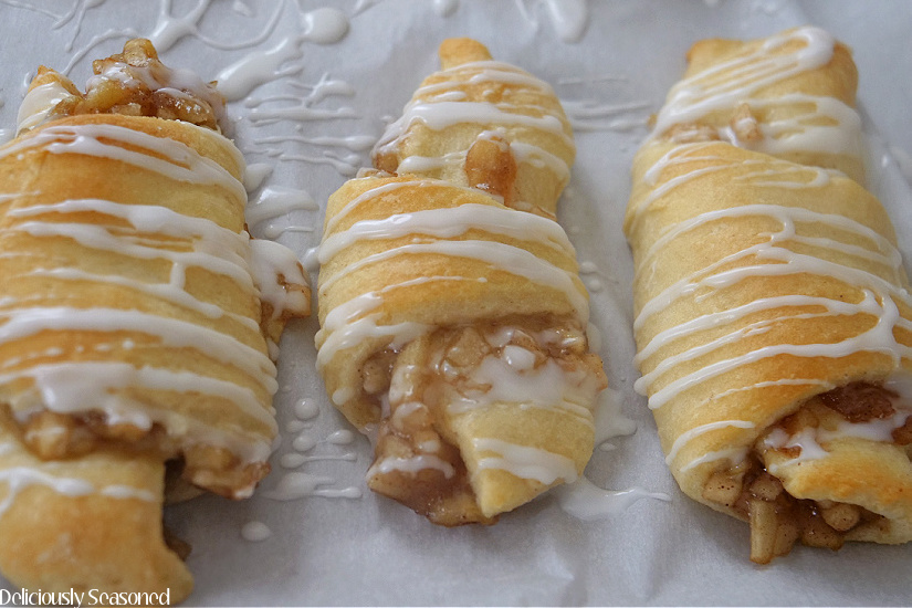Three crescents laying on parchment paper filled with apple pie filling and topped with glaze drizzle.