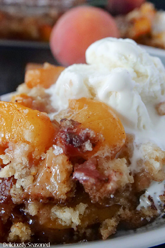 Peach cobbler in a white dish, topped with vanilla ice cream and a large peach in the background.