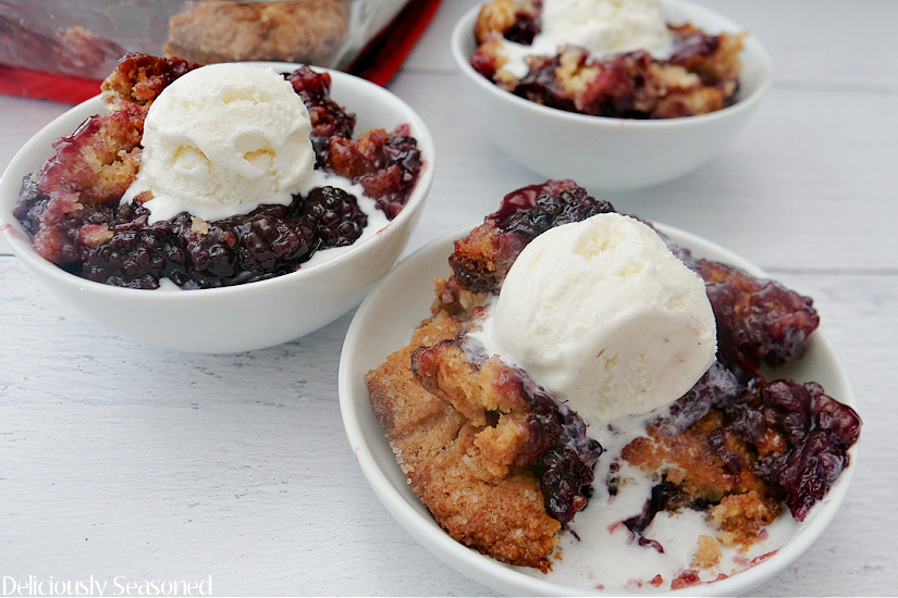 Three white bowls with blackberry cobbler in them all with scoops of vanilla ice cream on them.