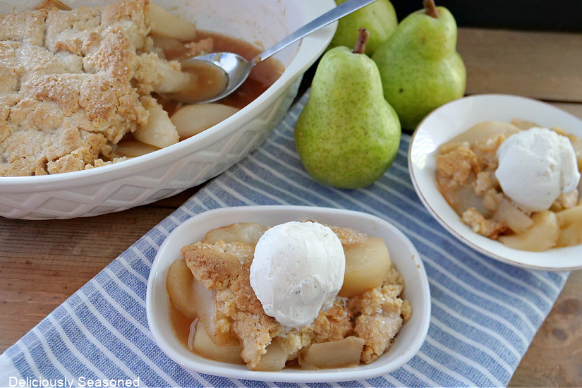 A oval baking dish with pear cobbler in it and two white bowls with servings of pear cobbler and three pears in the background.