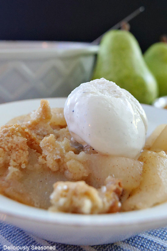 A close up photo of pear cobbler in a white bowl with a scoop of vanilla ice cream on top.