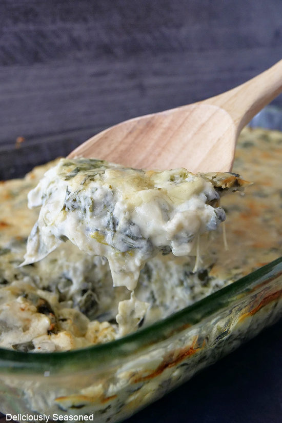 A glass baking dish filled with spinach artichoke dip and a wooden spoon spooning a scoop out.