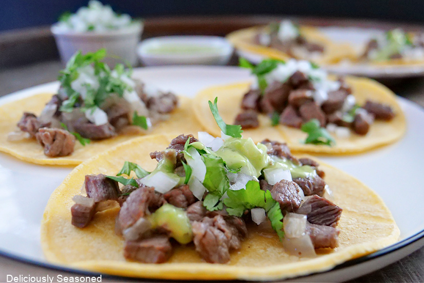 A large white plate with three corn tortillas laid out, with filling of beef, onions, cilantro, and a creamy green sauce on top.