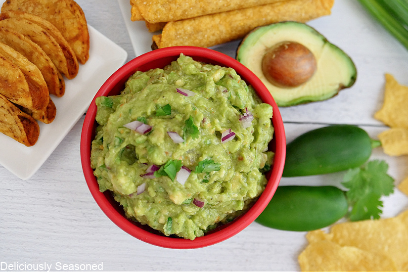 An overhead photo of a red bowl filled with guacamole with mini tacos on a white plate and whole jalapenos, tortilla chips and half an avocado also in the photo.