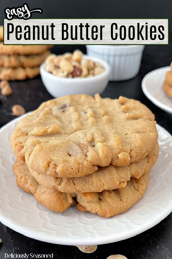 Three peanut butter cookies stacked up on a white plate with small bowls of peanut butter and chocolate chip morsels in the background.