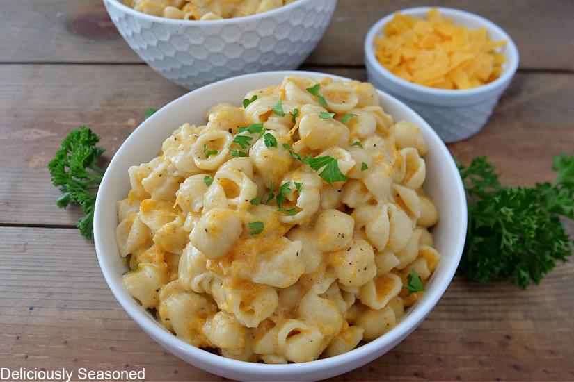 A white bowl filled with butternut squash mac and cheese with parsley and shredded cheese in the background for decoration.
