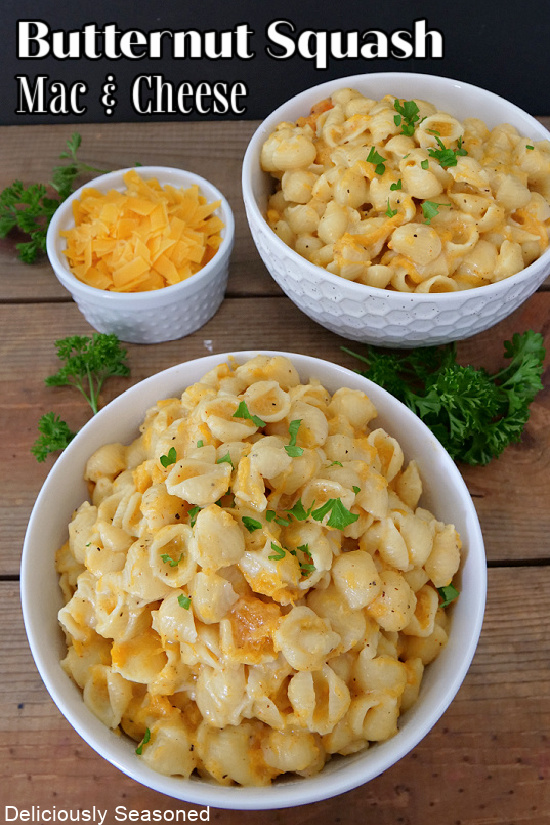 Two white bowls of macaroni and cheese with a small white bowl of shredded cheddar cheese also.