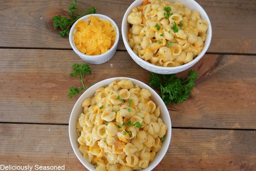 An overhead photo of a few bowls of mac and cheese with a small bowl of shredded cheese next to it, along with sprigs of parsley.
