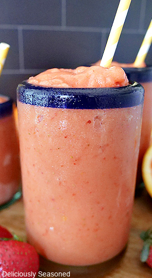 A close up picture of a Strawberry Peach Smoothies in a clear glass with blue trim with strawberries and oranges in the background.