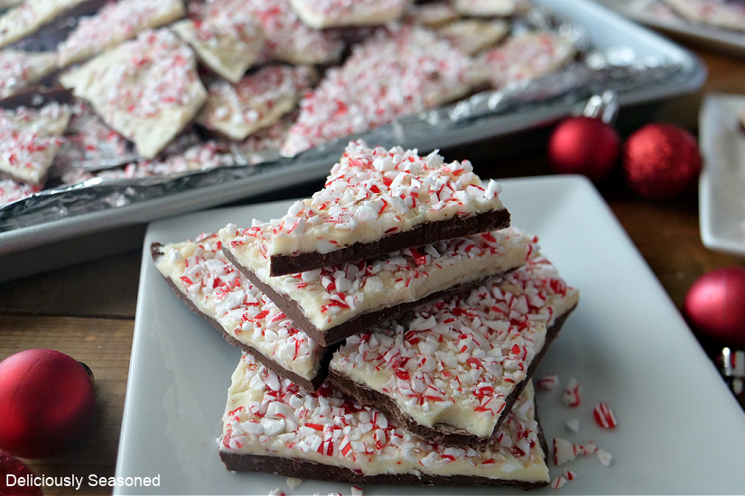 Peppermint bark stacked on a white plate with a baking sheet full of peppermint bark in the background.
