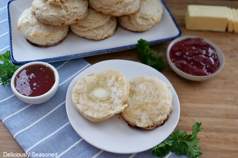 A biscuit cut in half, topped with melted butter. Two small white bowls filled with strawberry jelly in the background and a stack of biscuits in the back.