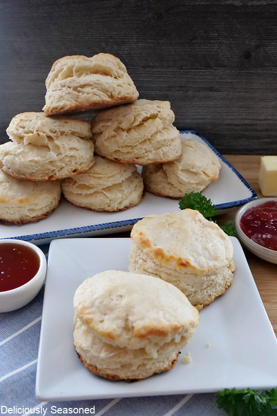 A small white square plate with two flaky biscuits sitting on it. A long plate in the back with biscuits stacked up, and two small bowls of strawberry jelly in the background.
