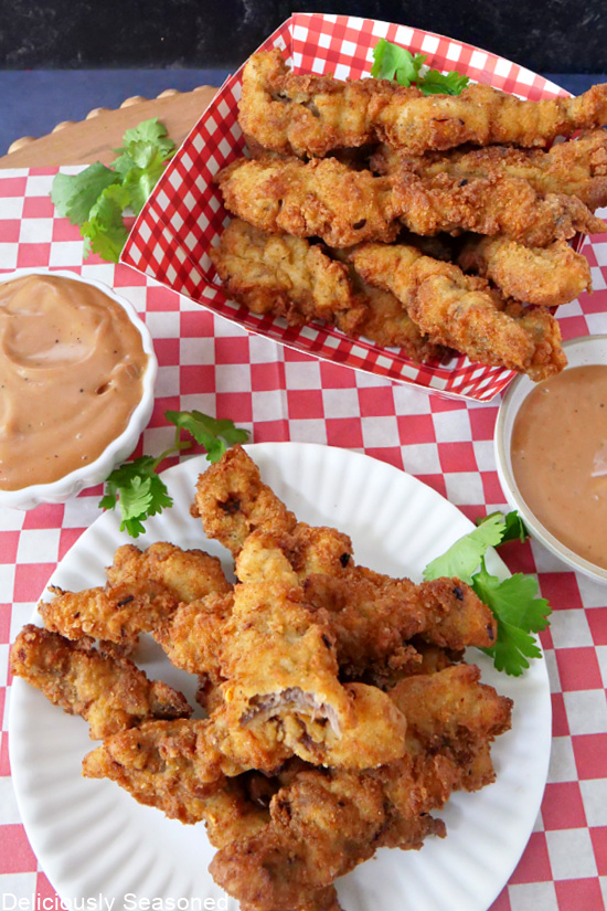 An overhead photo of a basket of fried steaks fingers, with some on a white plate, and two small white bowls filled with a dipping sauce.