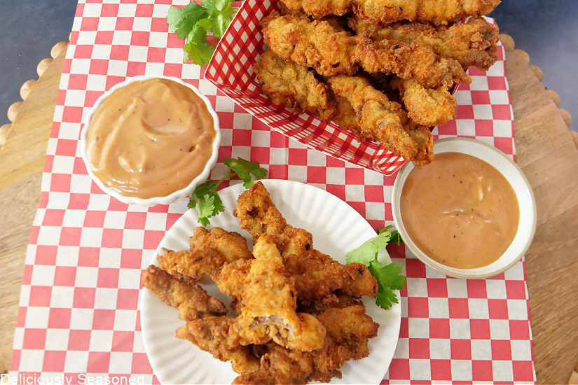 A red and white checkered board with a basket of steak fingers and a white plate loaded with them, with 2 small bowls of dipping sauce on it too. 