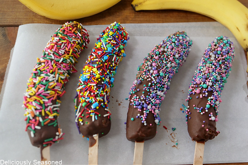 Four chocolate covered bananas covered in sprinkles, laying on parchment paper, with bananas in the background.