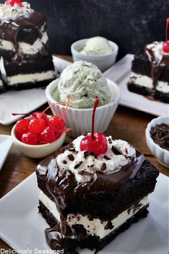 A few square, white plates with pieces of chocolate mint cake on them, all topped with hot fudge, whipped cream, and a cherry.