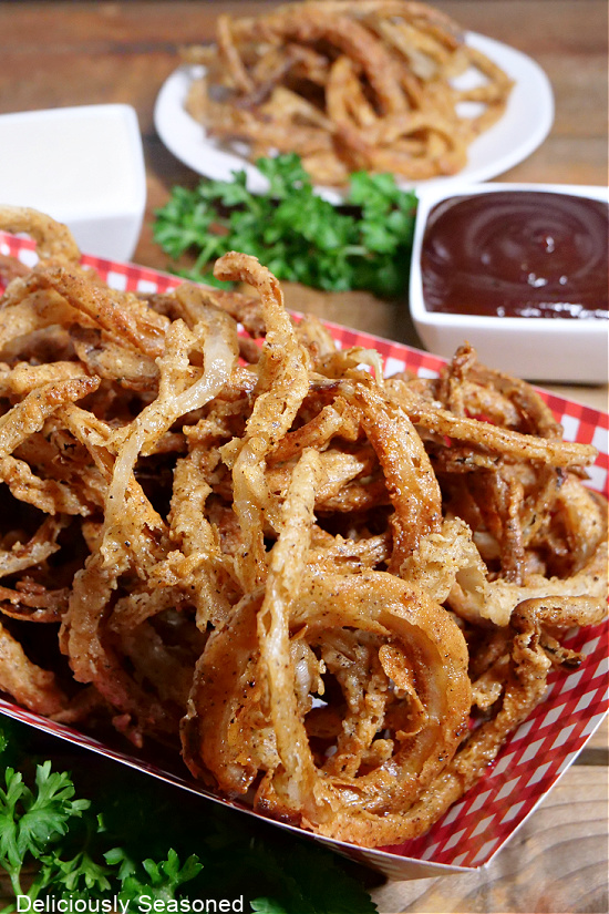 A red and white checkered basket filled with onion strings and a small bowl of BBQ sauce in the background.