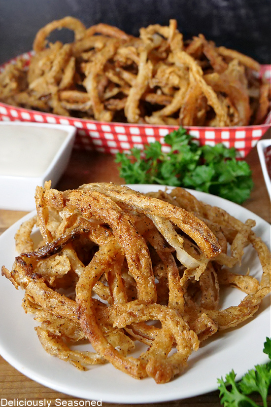 crispy onions strings on a round white plate with a red and white checkered basket in the background with a small bowl of ranch dressing. 