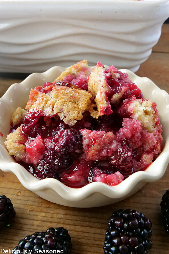 A white bowl filled with blackberry cobbler with blackberries in the foreground.