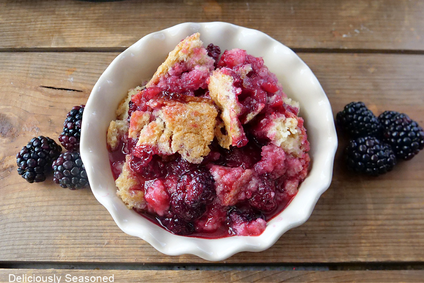 A horizontal photo of blackberry cobbler in a white bowl surrounded by fresh blackberries.