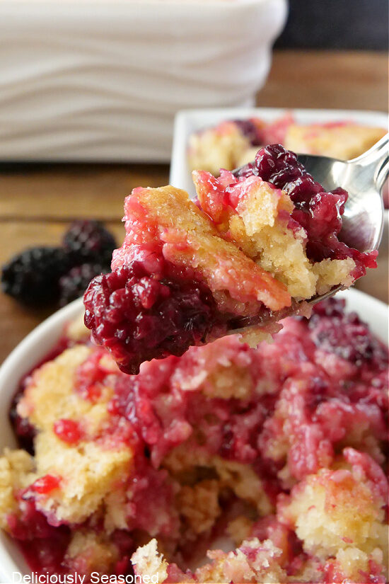 A close-up picture of a spoonful of blackberry cobbler over a bowl filled with blackberry cobbler and blackberries in the background.