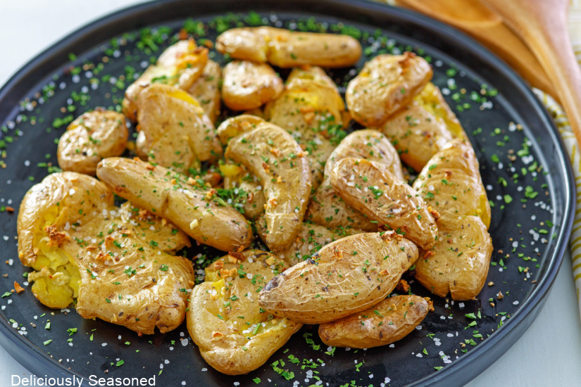 A black plate with smashed baby potatoes, roasted garlic, salt and pepper and parsley on them.
