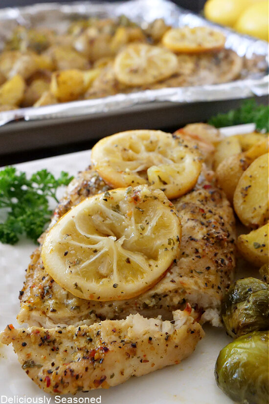 Lemon pepper chicken on a white plate topped with lemon slices and surrounded with Brussel sprouts and potatoes.