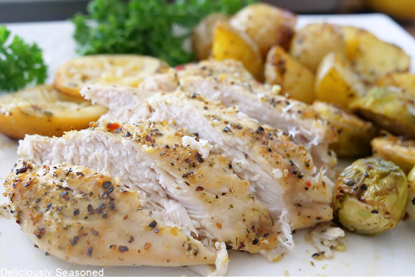 A horizontal picture of a sliced chicken breast on a white plate with potatoes, Brussel sprouts, parsley, and lemon slices.